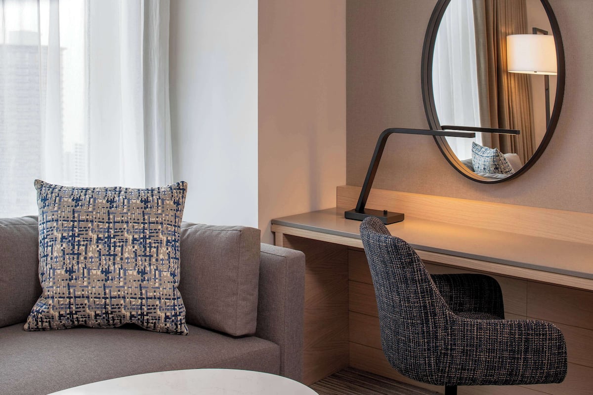 A modern hotel room with a grey sofa, a patterned pillow, a round mirror, and a dark armchair.