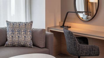 Una habitación de hotel moderna con un sofá gris, una almohada estampada, un espejo redondo y un sillón oscuro.
