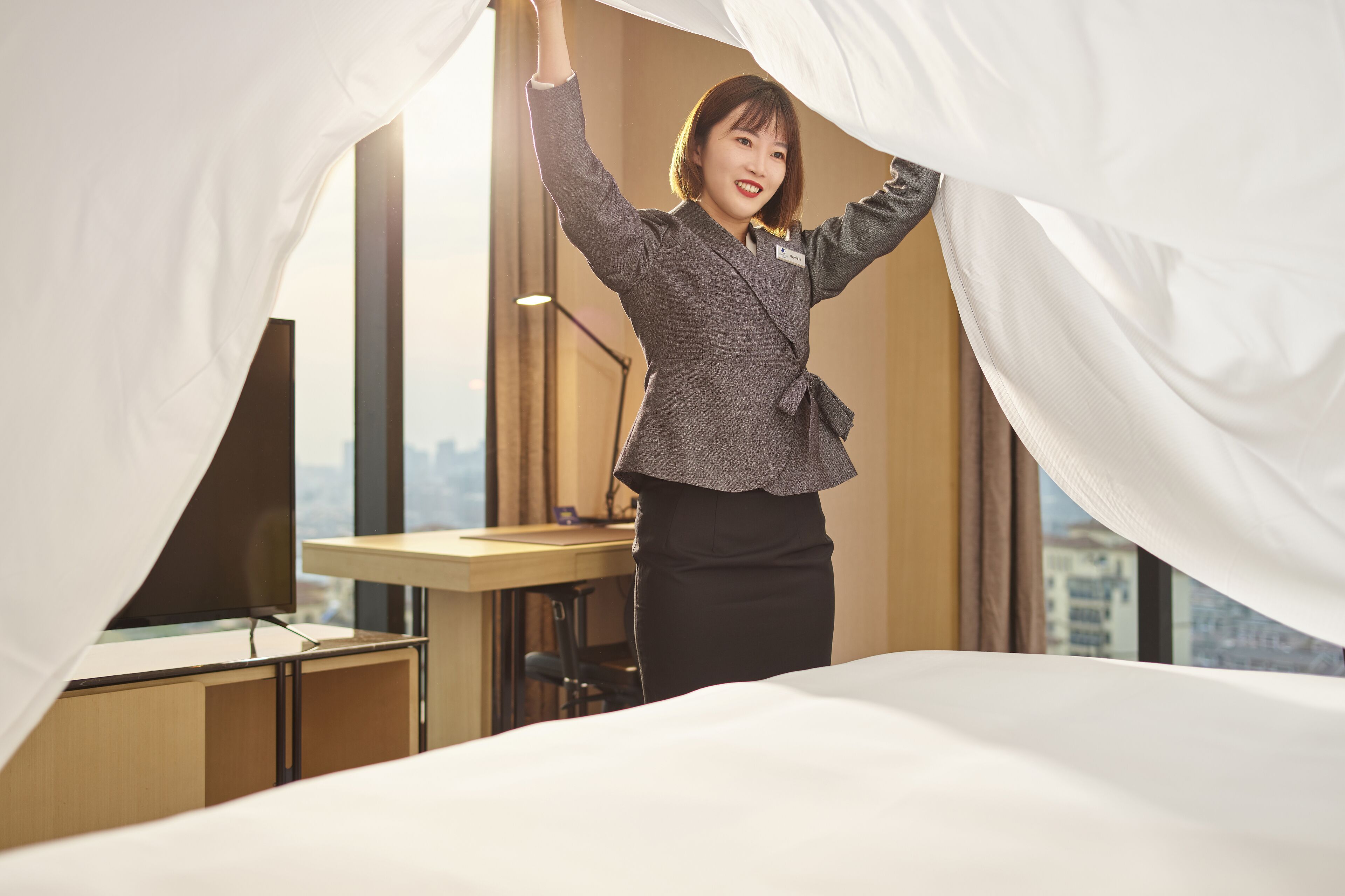 Habitación de hotel con cama, escritorio y vistas a la ciudad a través de amplias ventanas.