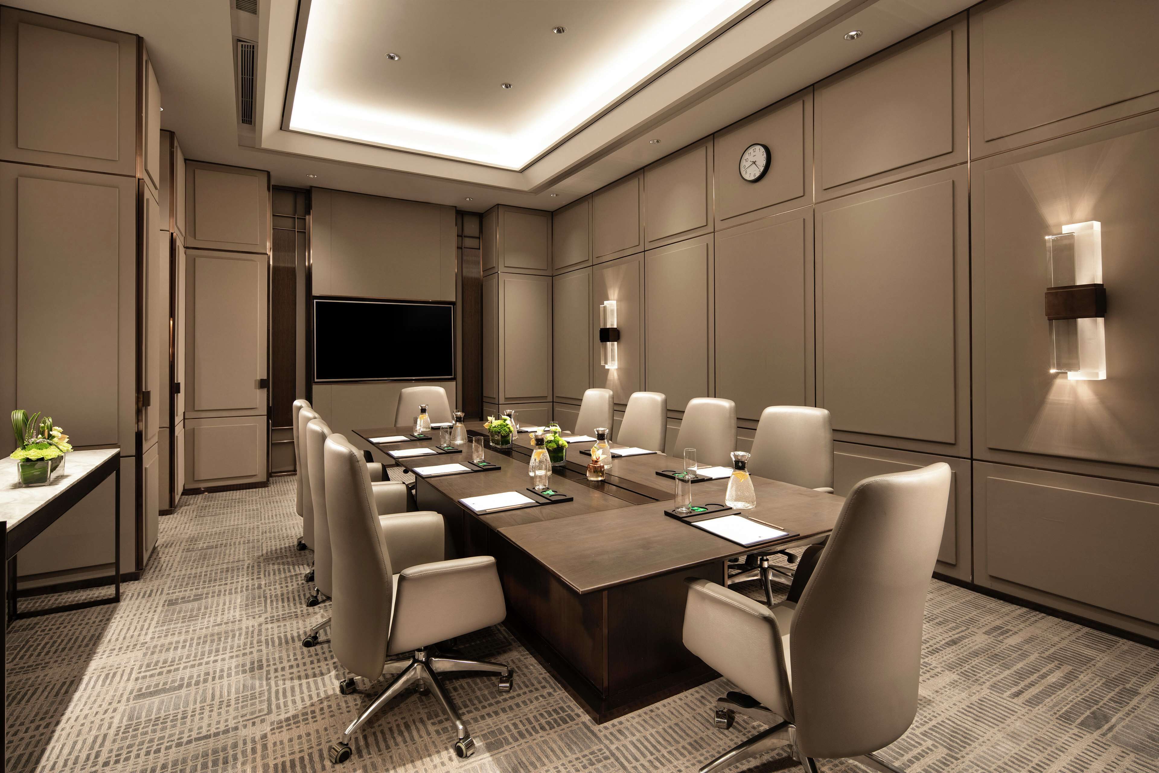 A conference room with a long table, chairs, a flat-screen TV, and wall-mounted lighting.