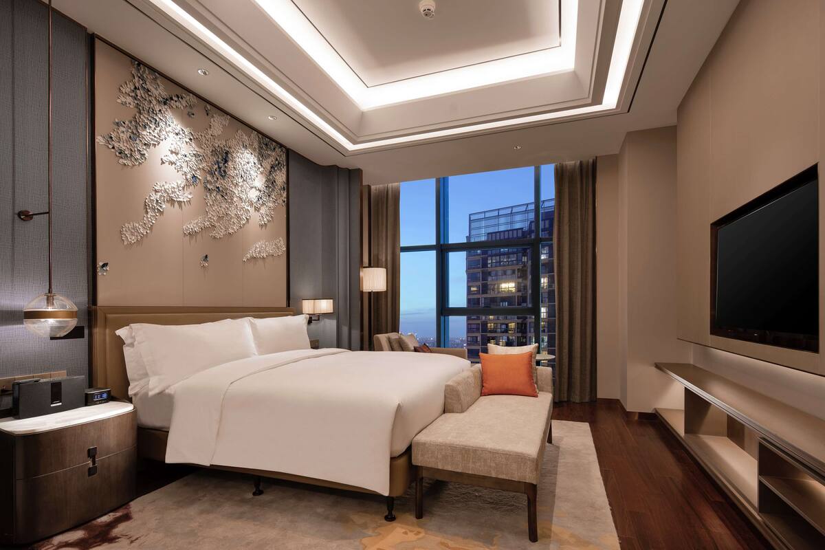 A modern hotel room with a large bed, a flat-screen TV, a desk, and a view of a cityscape through large windows.