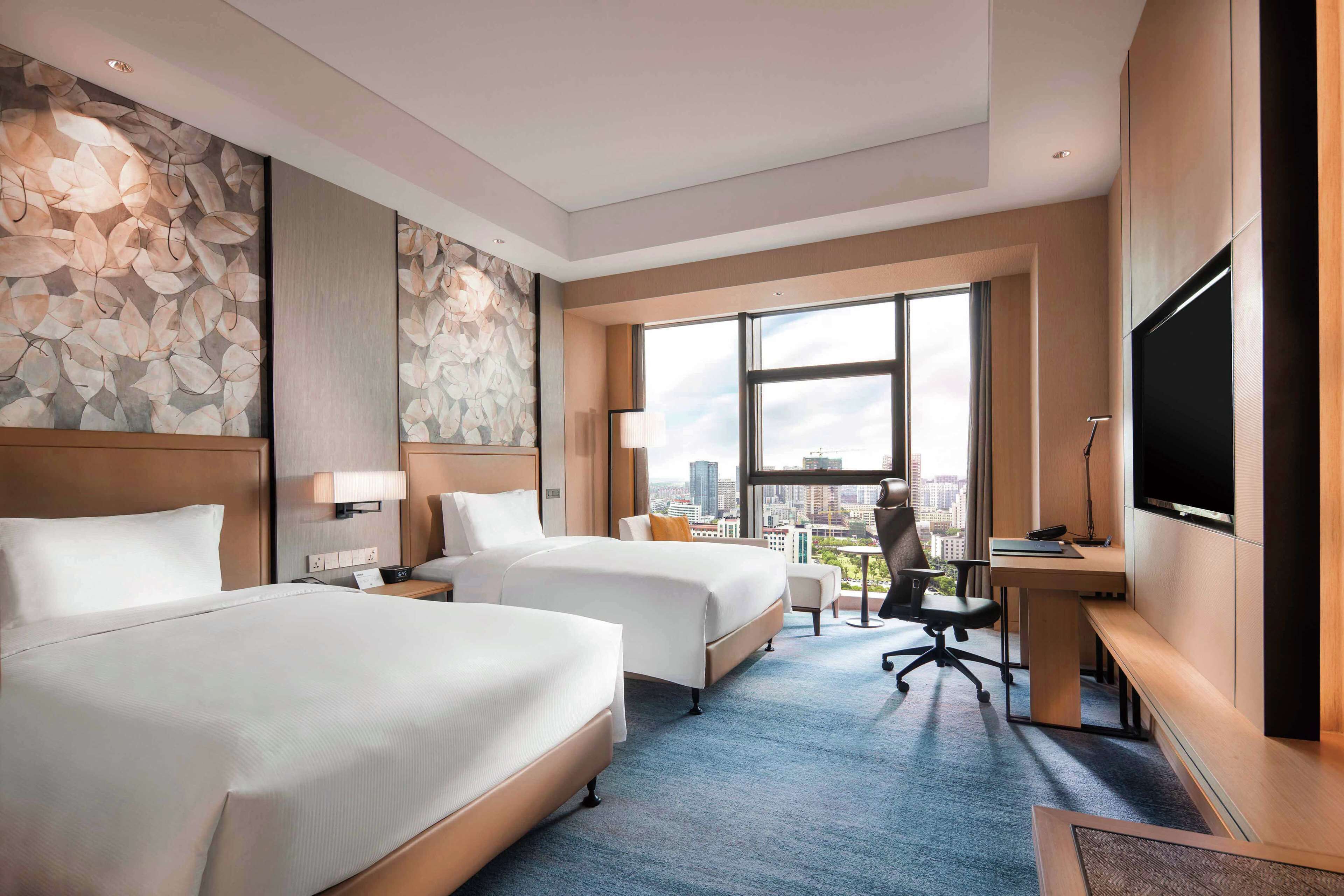 A hotel room with two beds, a desk, a TV, and a city view.