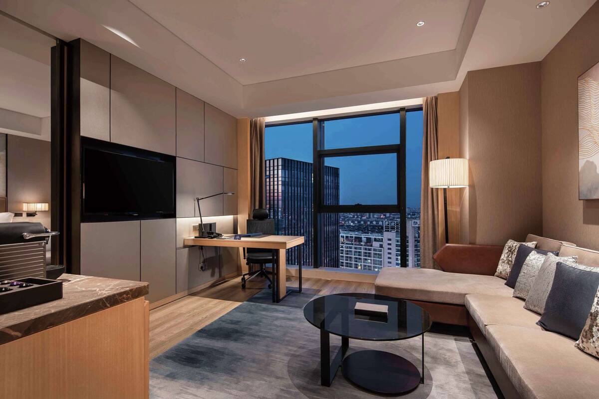 A modern hotel room with a large TV, a desk, a sofa, and a view of the cityscape.