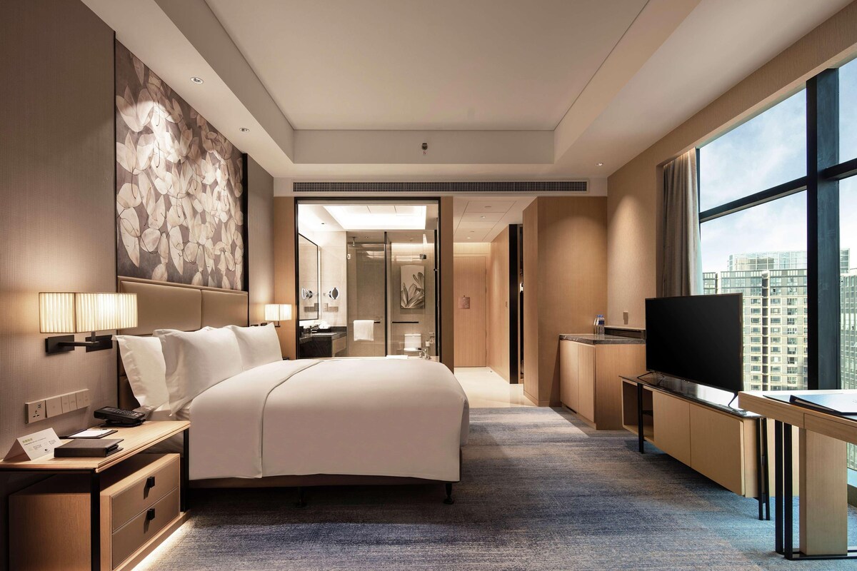 A modern hotel room with a large bed, a desk with a flat-screen TV, a large window with a city view, and a bathroom visible through a glass door.