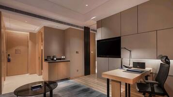 Una habitación de hotel moderna con un escritorio de madera, una silla de oficina negra, un televisor de pantalla plana montado en la pared y una pequeña zona de kitchenette.
