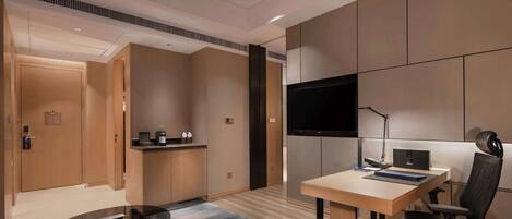 Una habitación de hotel moderna con un escritorio de madera, una silla de oficina negra, un televisor de pantalla plana montado en la pared y una pequeña zona de kitchenette.