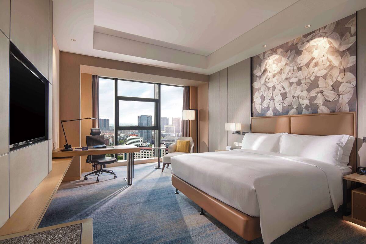 A modern hotel room with a large bed, a desk with a chair, a flat-screen TV, and a city view through large windows.