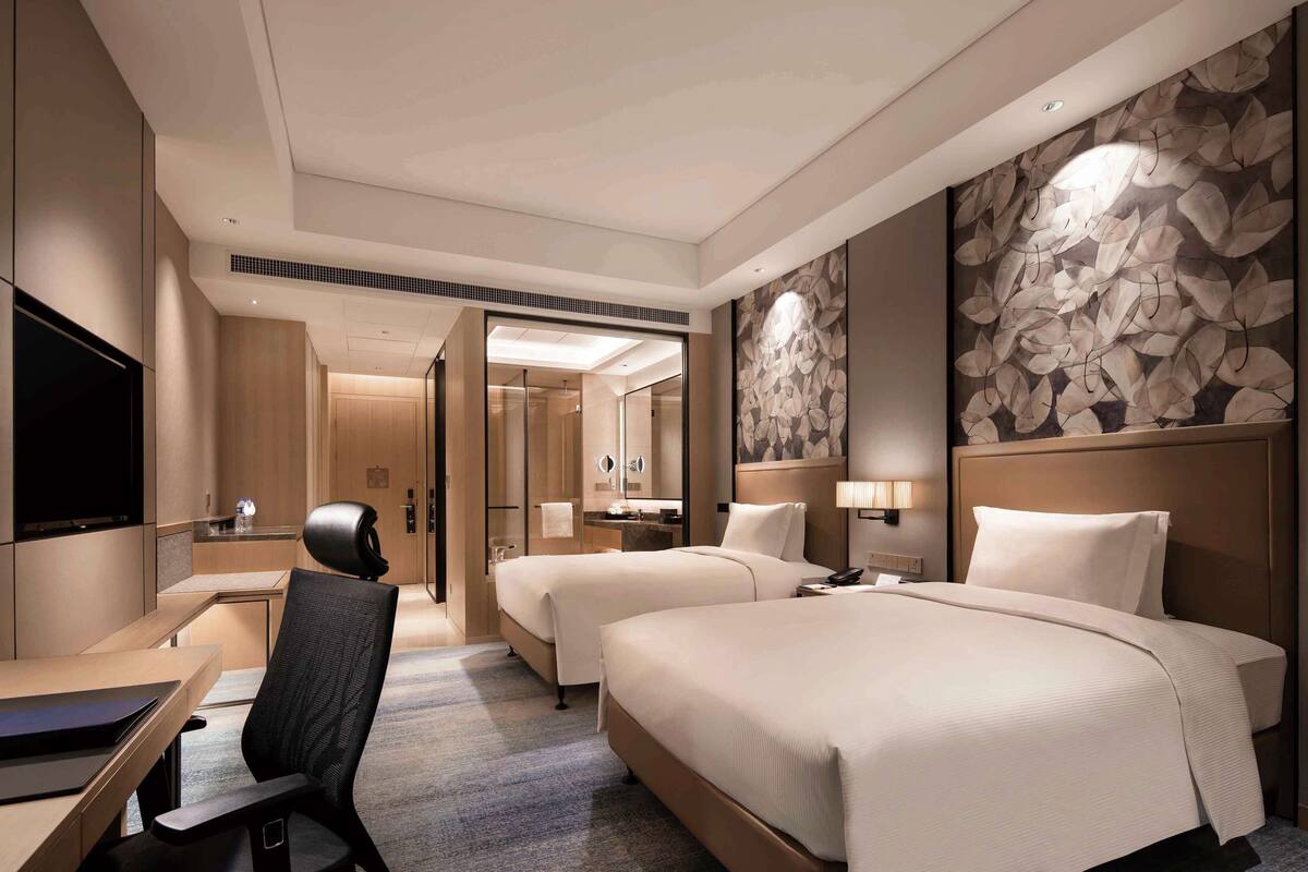 A hotel room with two beds, a desk, a chair, a TV, and a bathroom visible through an open door.