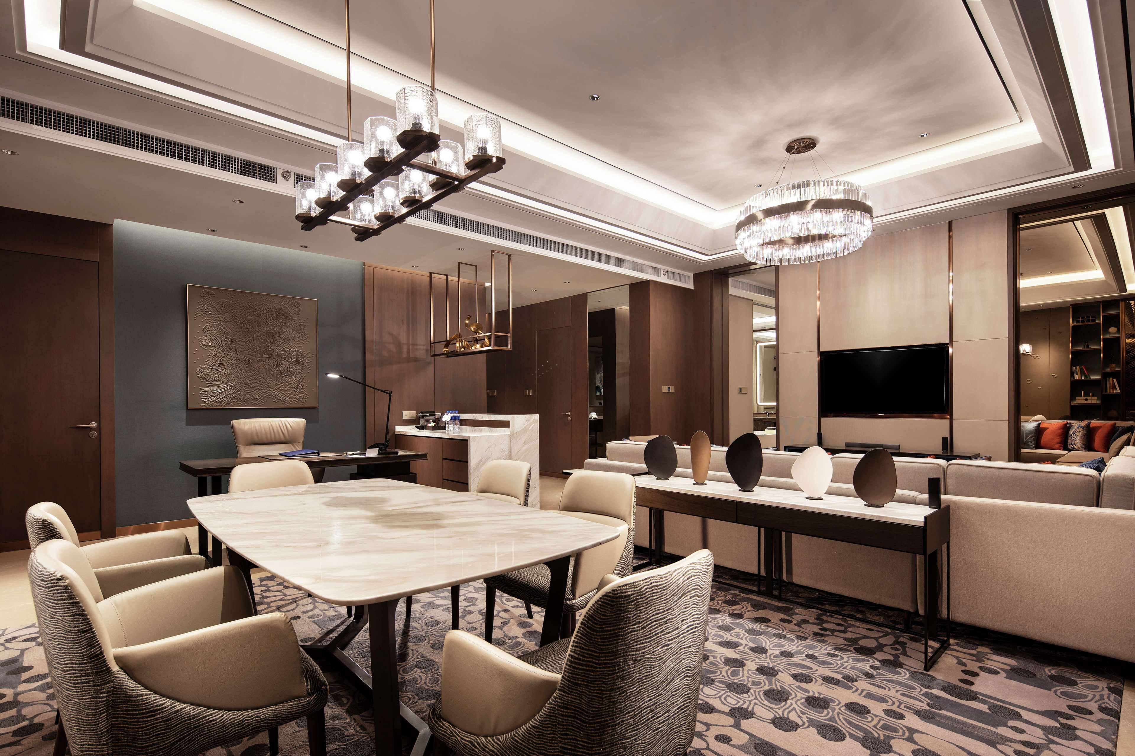 A modern hotel room with a dining area, a sofa set, a flat-screen TV, and a chandelier.