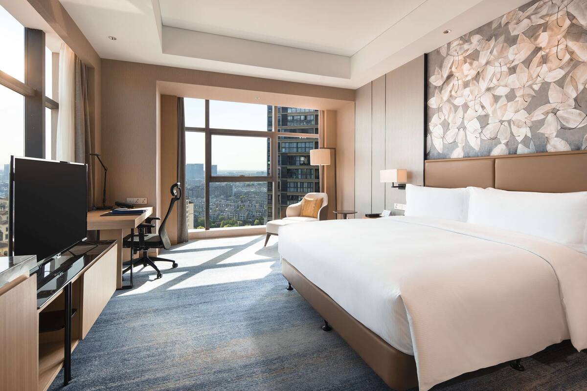 A hotel room with a large bed, a desk with a computer, a chair, a TV, and a large window with a city view.
