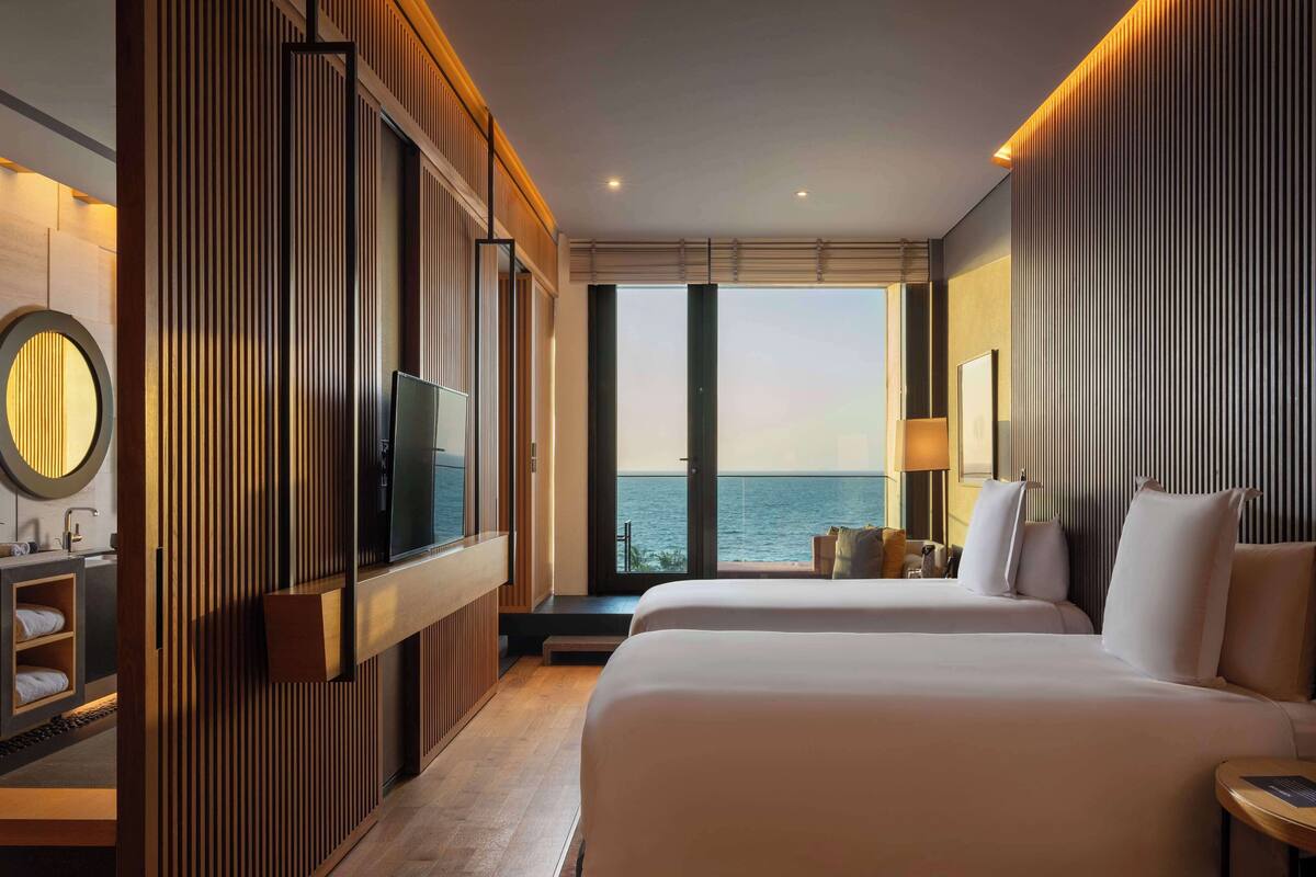 A hotel room with a large bed, a TV mounted on a wooden panel, a bedside table, and a view of the sea.