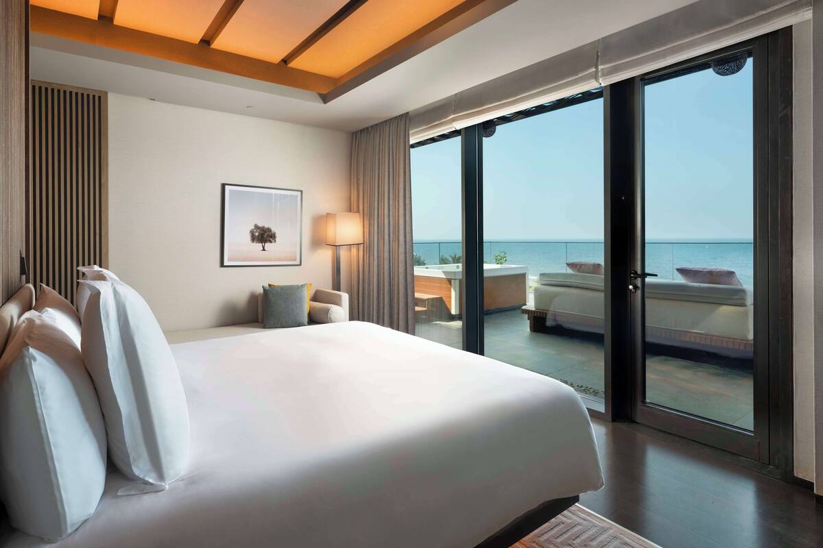 A hotel room with a large bed, a desk, a chair, and a view of the sea through a sliding door.
