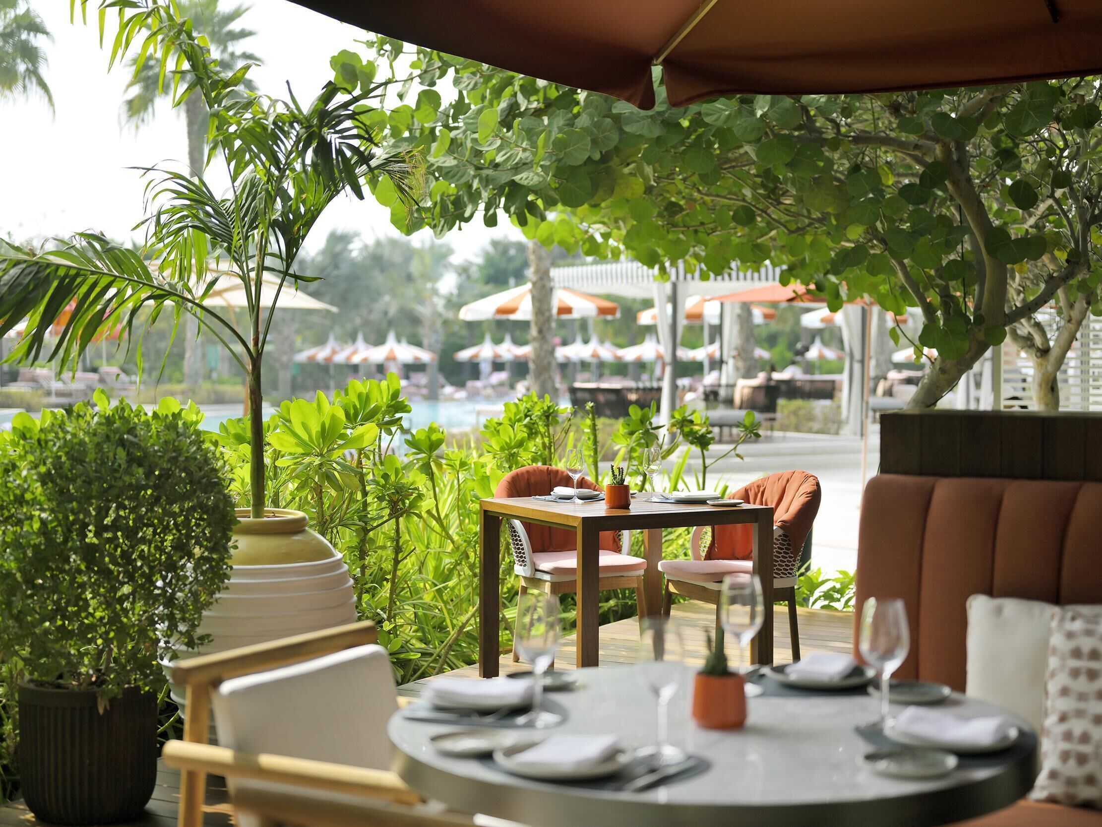 An outdoor dining area with a table set for two, surrounded by lush greenery and overlooking a pool area.