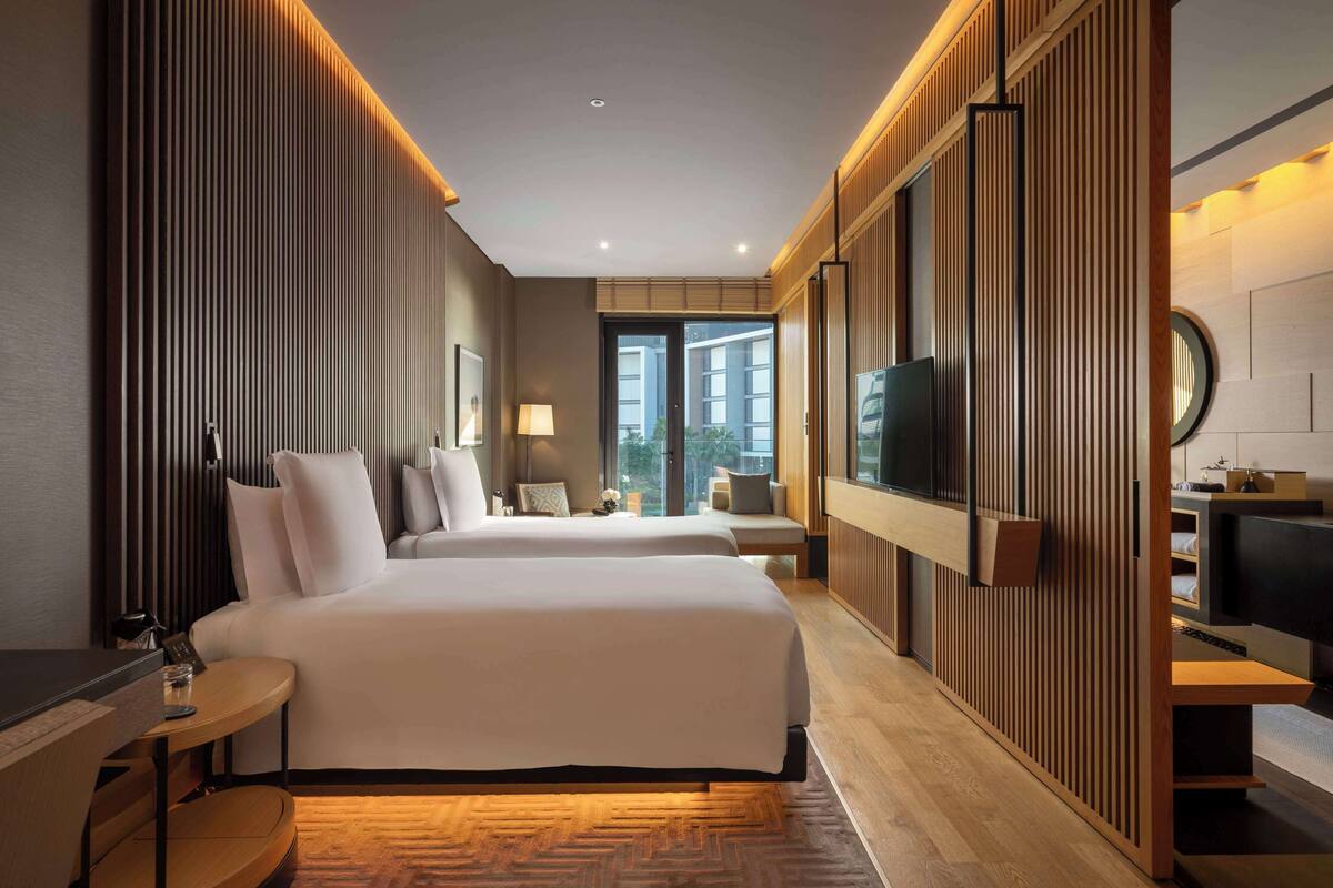 A modern hotel room with two beds, a TV, a desk, and a large window offering a city view.