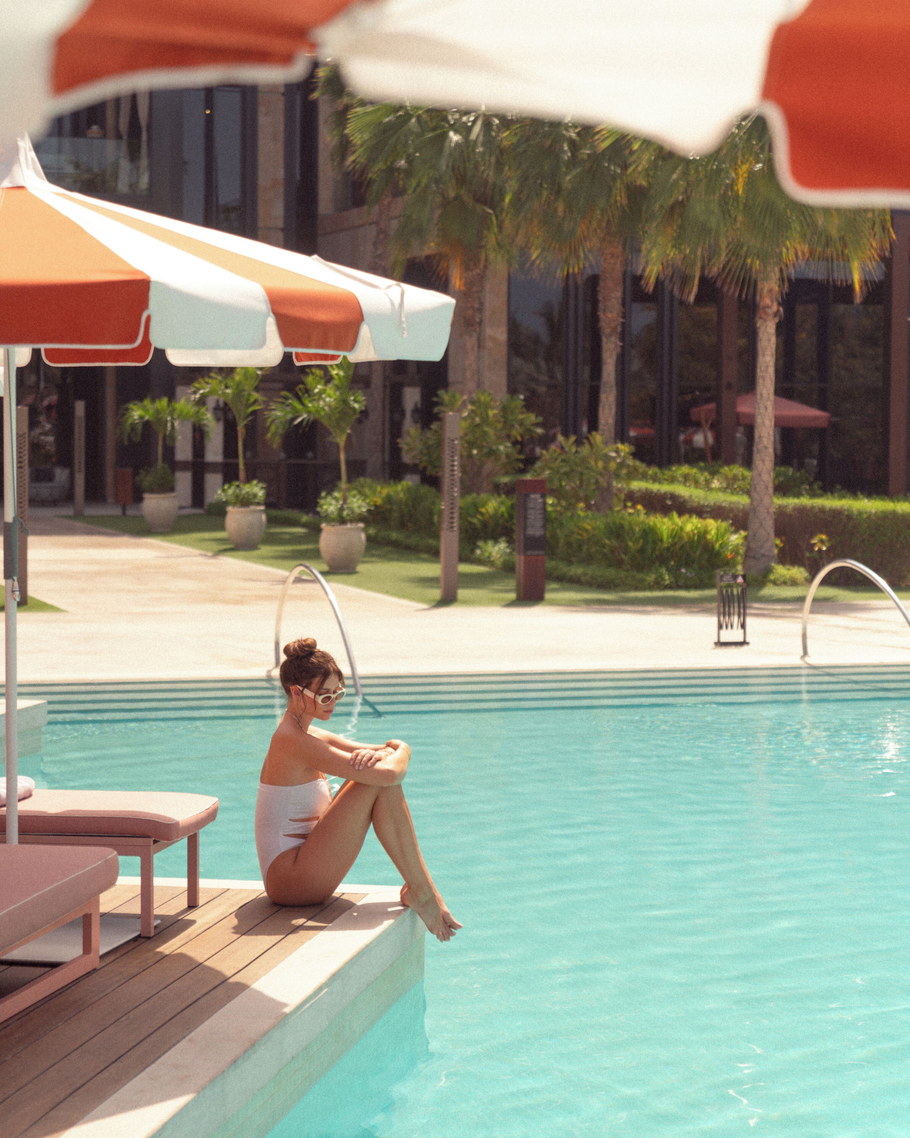 A poolside area with a person sitting by the pool, a lounge chair, and a palm tree in the background.