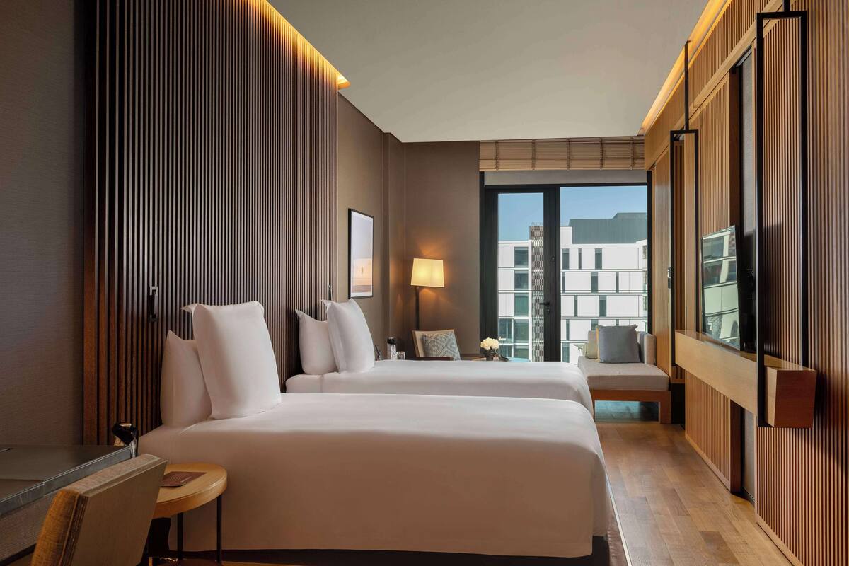 A hotel room with two beds, a desk, a chair, a lamp, and a view of buildings through the window.