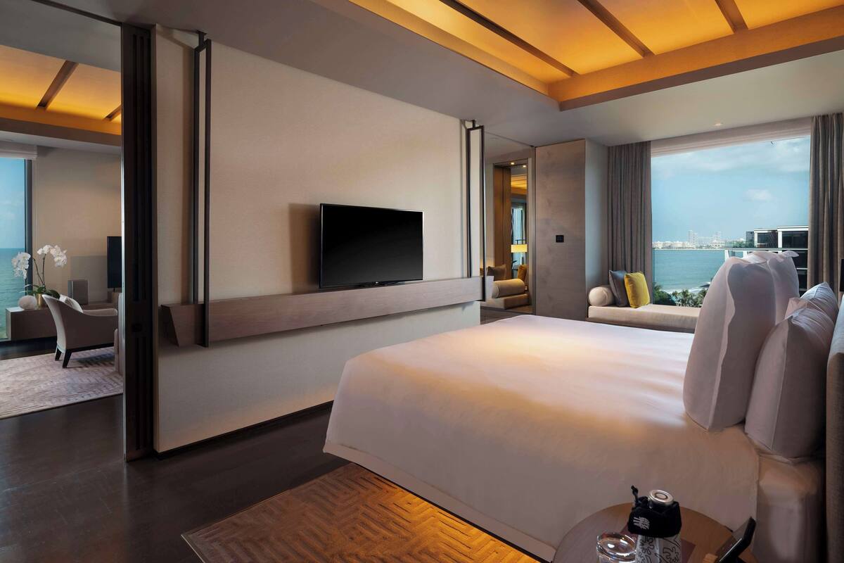 A modern hotel room with a large bed, a flat-screen TV mounted on the wall, a seating area with a sofa and armchairs, and a view of the sea through large windows.