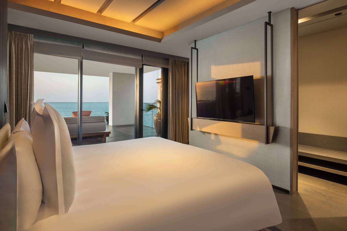 A modern hotel room with a large bed, a flat-screen TV mounted on the wall, and a balcony with a view of the sea.