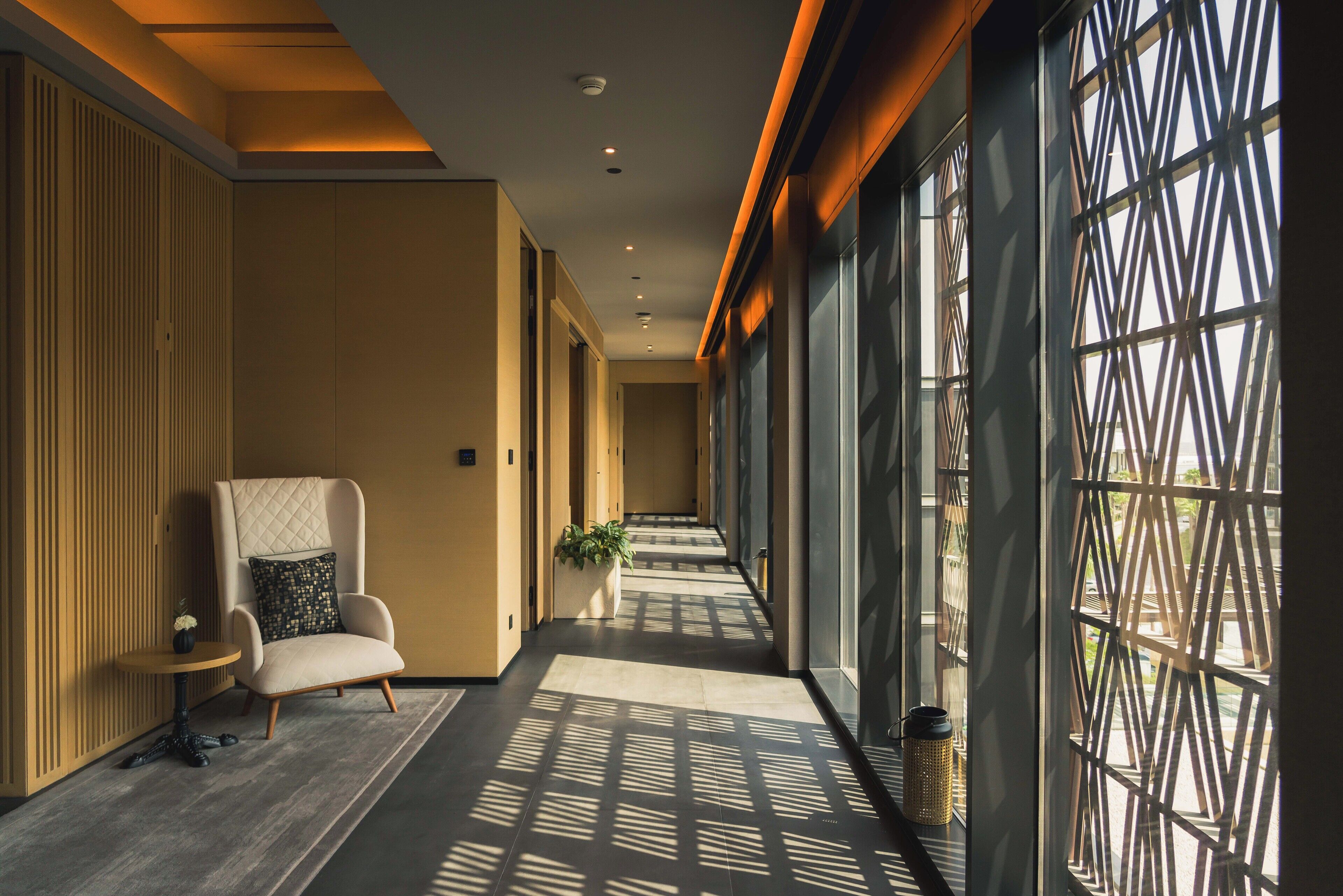 A hallway with a modern design, featuring a seating area with a chair and a small table, a potted plant, and a decorative wall with a geometric pattern.