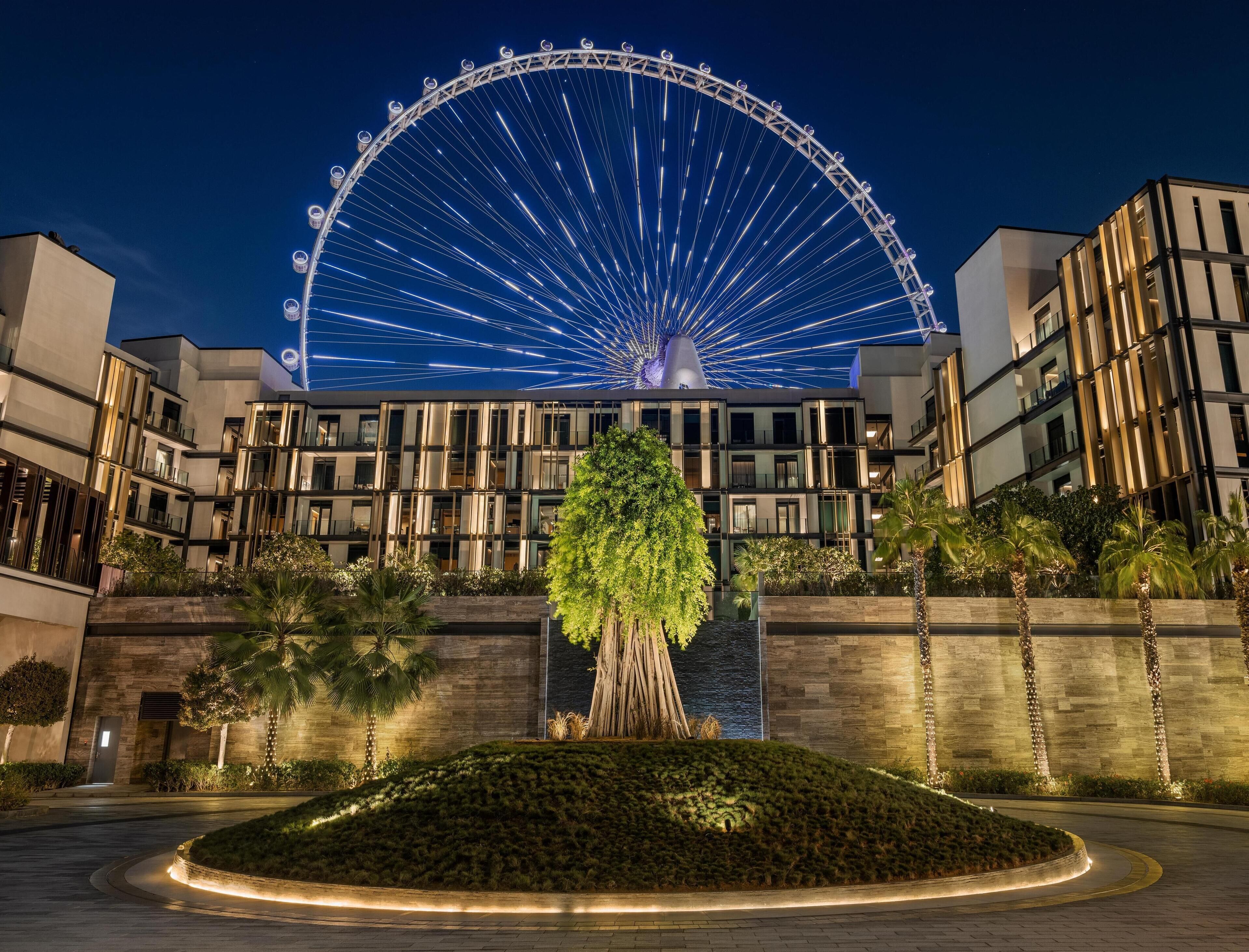 A modern building with a large Ferris wheel in the background, surrounded by landscaped gardens and palm trees.
