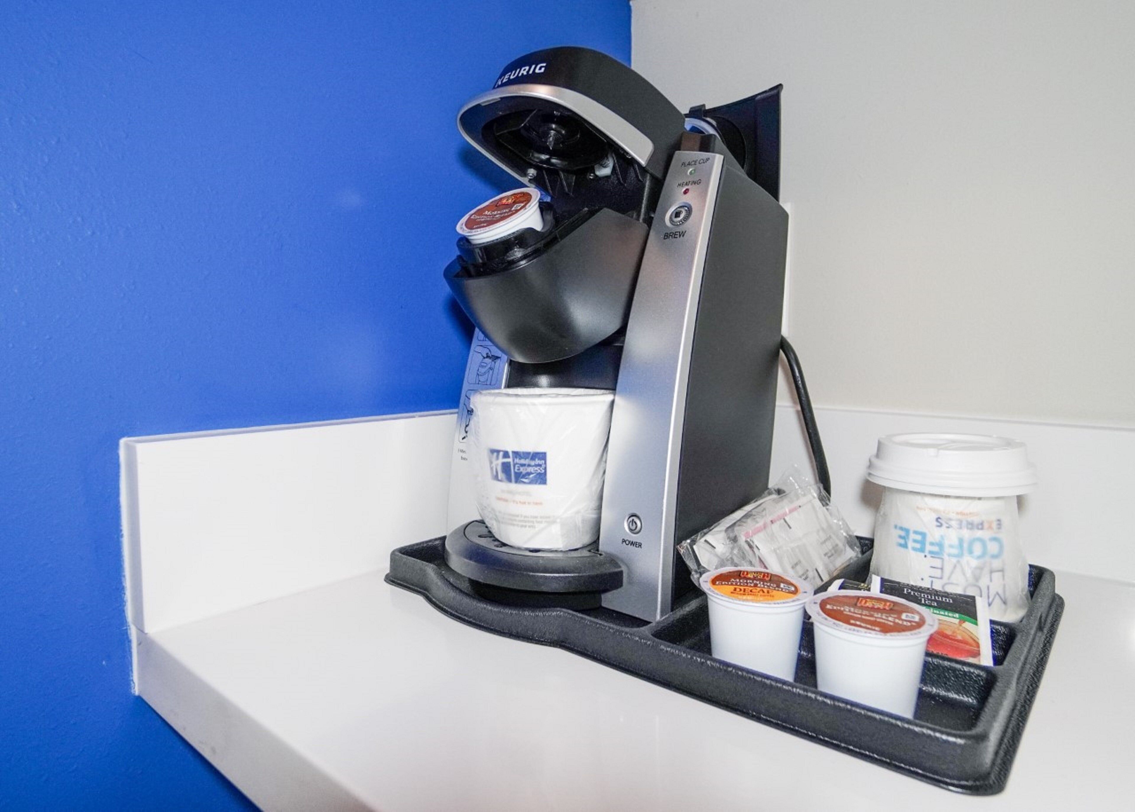 Una cafetera con una taza de café y dos recipientes de crema sobre una encimera blanca frente a una pared azul.