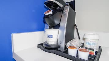 Una cafetera con una taza de café y dos recipientes de crema sobre una encimera blanca frente a una pared azul.