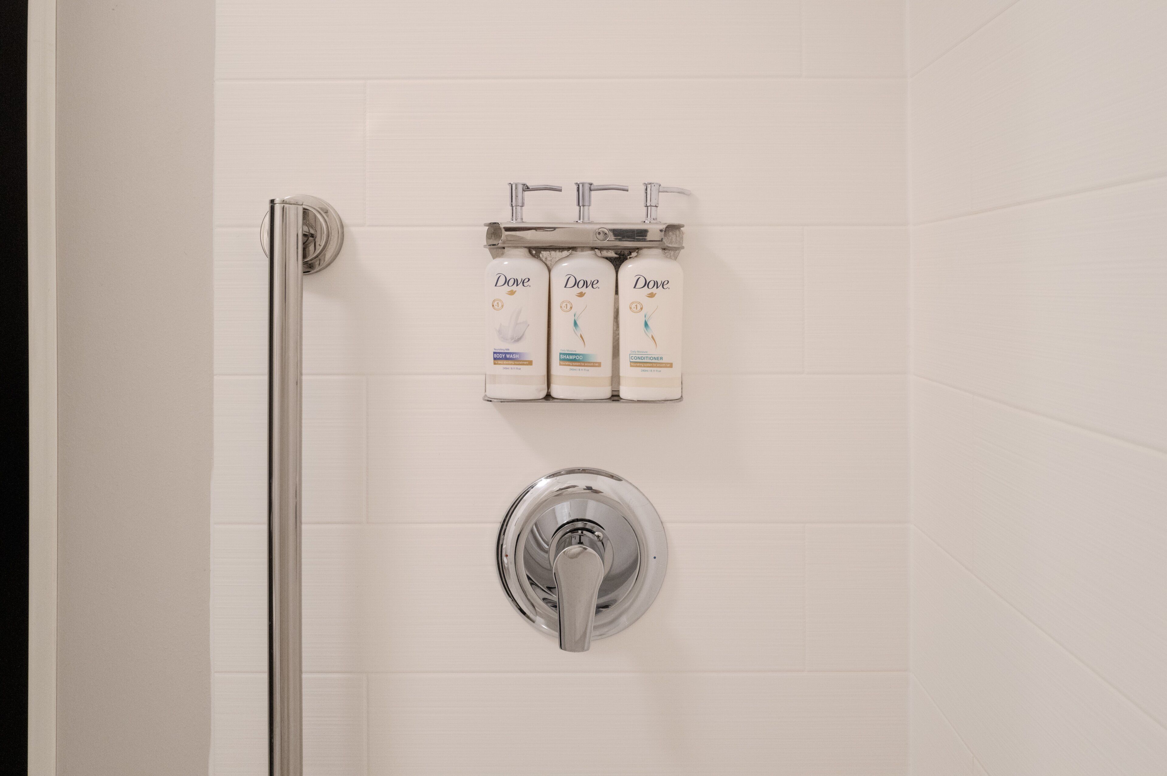 A bathroom with a white tiled wall, a chrome showerhead, and three Dove products on a shelf.