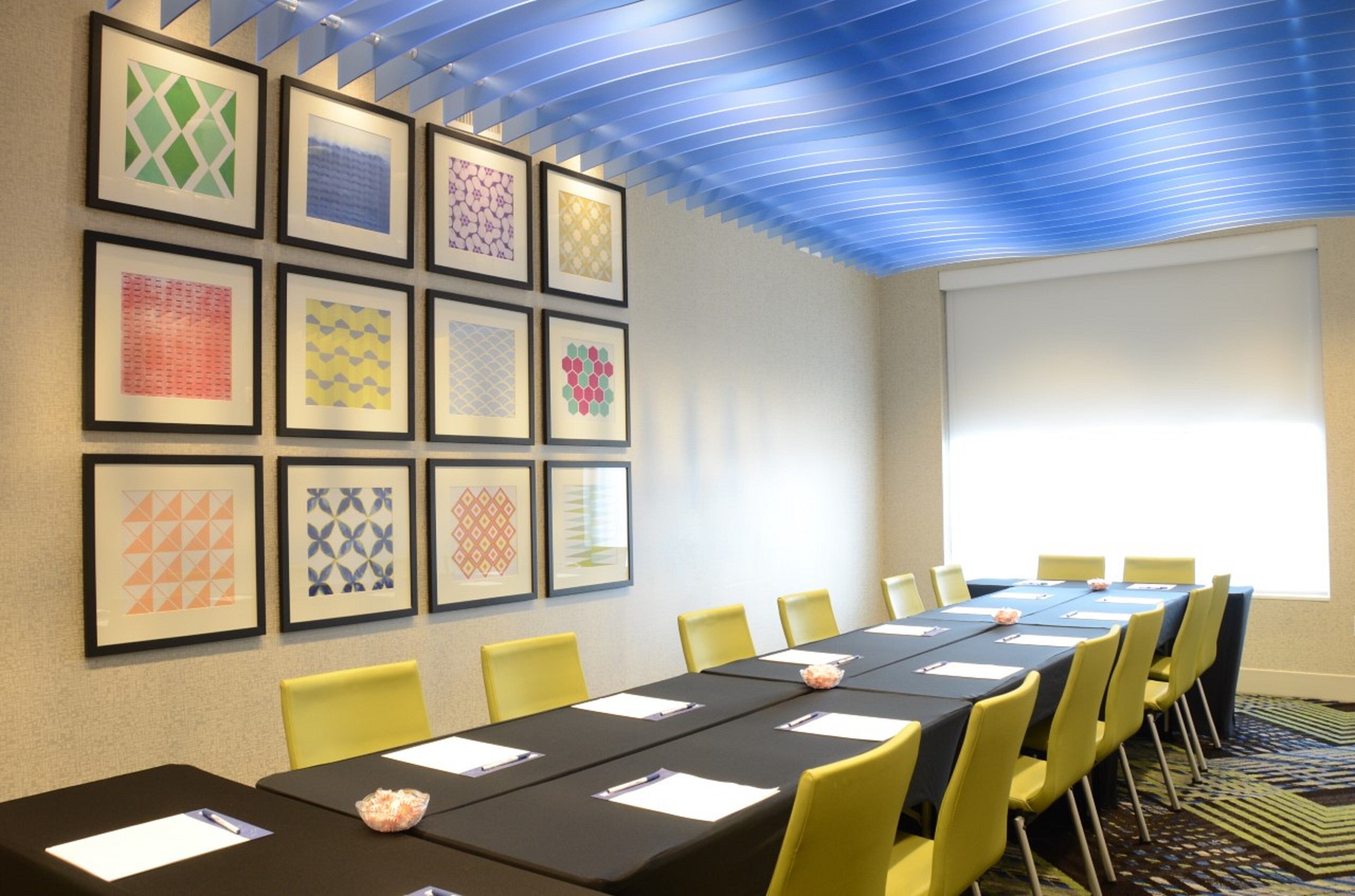 Une salle de conférence avec une longue table, des chaises jaunes, des motifs géométriques encadrés sur le mur et une grande fenêtre.
