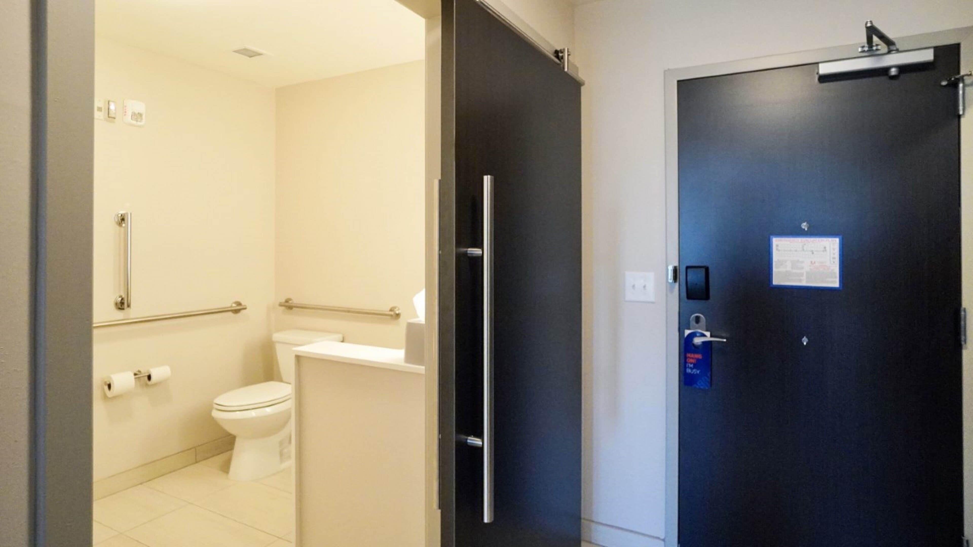 A bathroom with a toilet, handrails, and a towel dispenser.