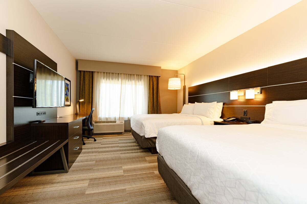 A hotel room with two beds, a desk with a flat-screen TV, a lamp, and a window with curtains.