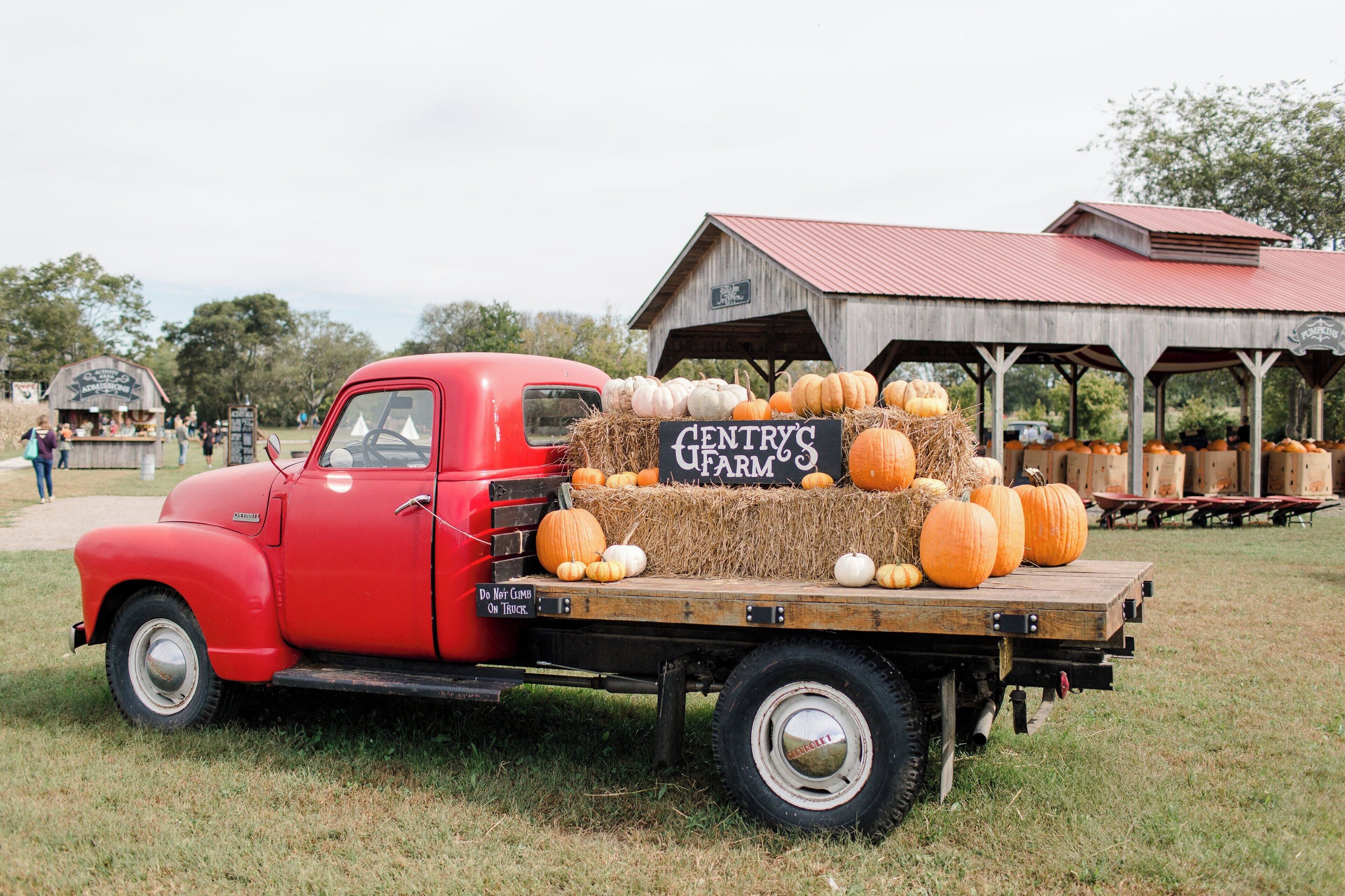 A vintage red truck with hay bales and pumpkins on the back, parked in front of a rustic barn with a sign that reads "CENTRY'S FARM."