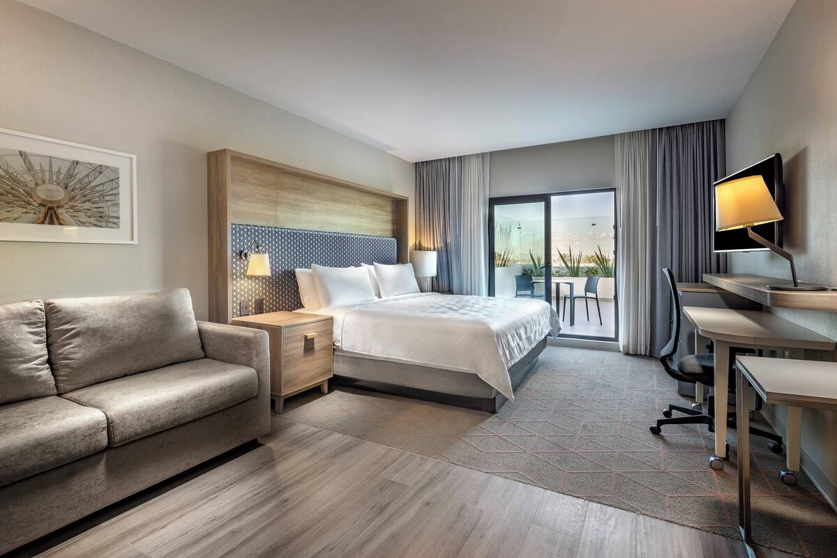 A modern hotel room with a bed, a sofa, a desk, a TV, and a balcony with curtains.