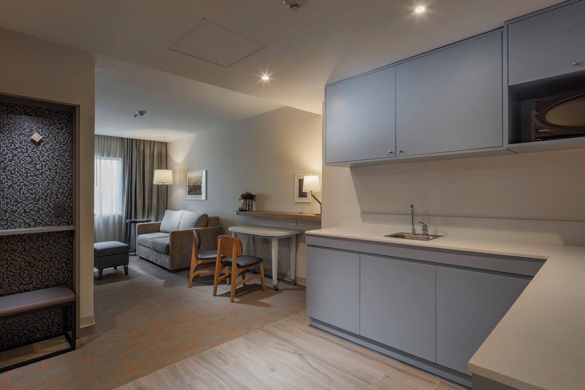 A modern hotel room with a kitchenette, a seating area with a sofa and chairs, and a patterned rug.