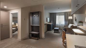 Una habitación de hotel moderna con kitchenette, zona de estar con sofá y sillones, televisión de pantalla plana y un ventanal con cortinas.