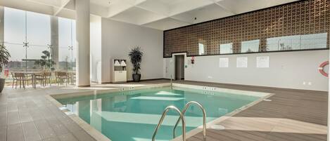 A modern indoor swimming pool with clear water, a tiled deck, and a view of the city through large windows.