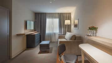 Ein Hotelzimmer mit Flachbildfernseher, einer Couch, einem kleinen Tisch, einem Sessel, einem Schreibtisch, einer Lampe und einem fenster mit Vorhängen.