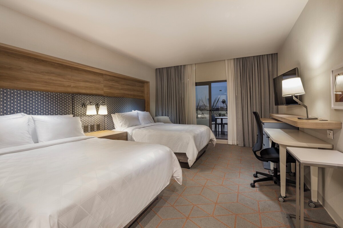 A hotel room with two beds, a desk, a chair, a TV, and a window with curtains.