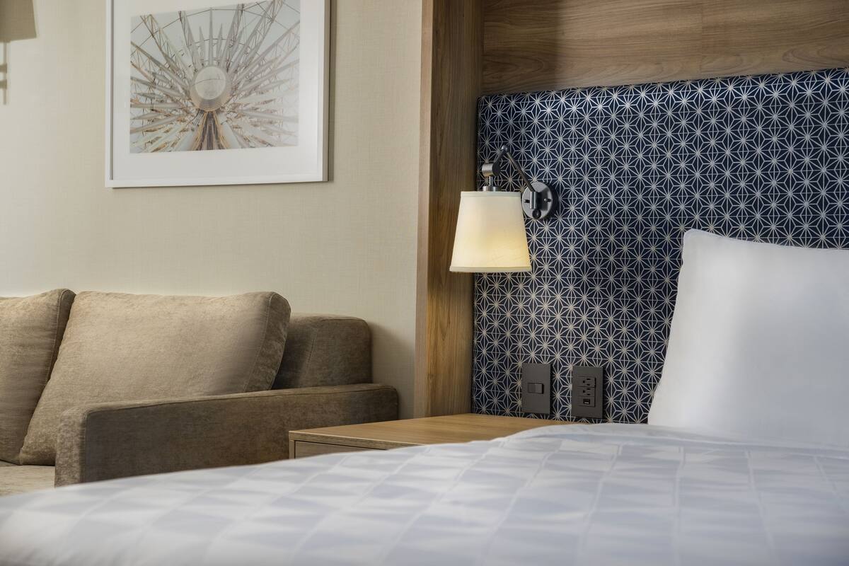 A hotel room with a bed, a nightstand, a lamp, a wall-mounted outlet, and a framed picture.