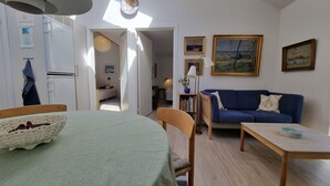 A living room with a blue sofa, a wooden coffee table, and a dining area with a table and chairs. There are paintings on the wall and a ceiling fan. - Penalhuzet (Ronne)