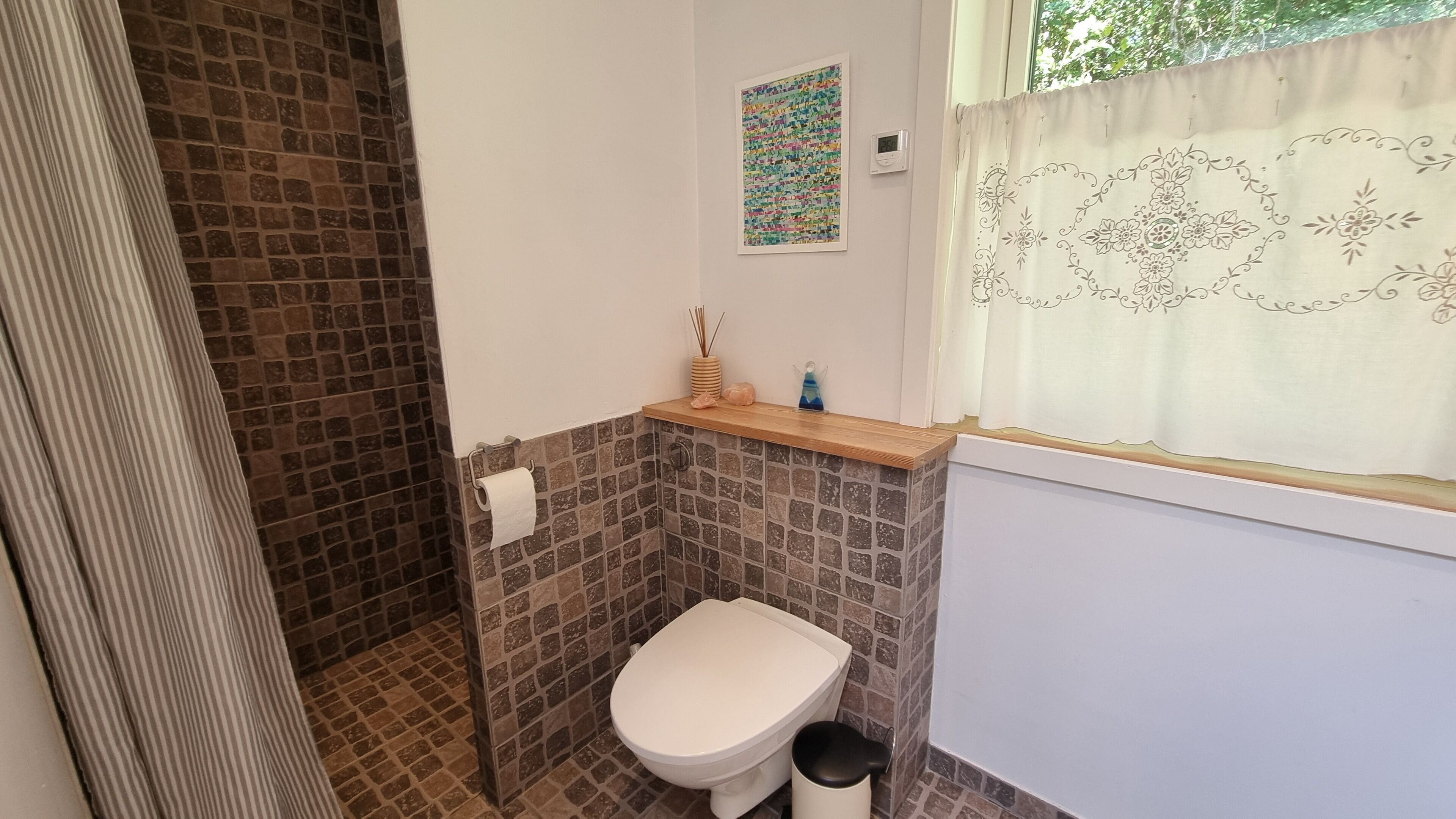 A small bathroom with a toilet, a shelf with toiletries, a decorative wall piece, and a window with curtains.