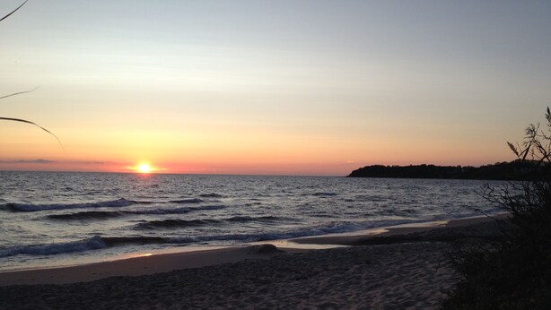 A beach with the sun setting over the ocean, a sandy shore, and some vegetation on the right. - Penalhuzet (Ronne)