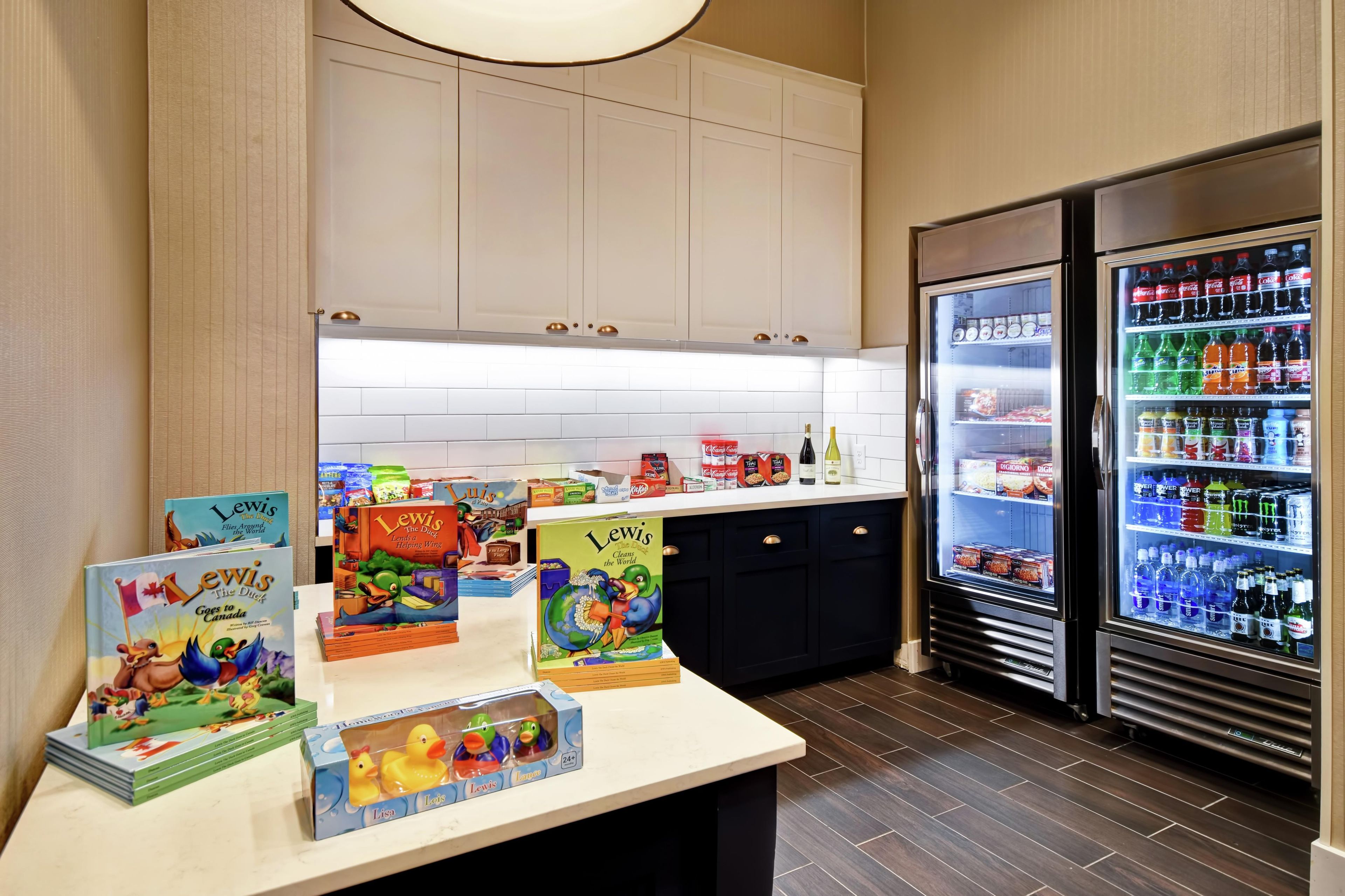 A hotel kitchen with a refrigerator stocked with beverages, a counter with children's books and toys, and cabinets above.