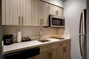 A modern kitchen with wooden cabinets, a stainless steel refrigerator, a microwave, a sink, and a coffee maker. - Homewood Suites by Hilton Greenville Downtown (Greenville)