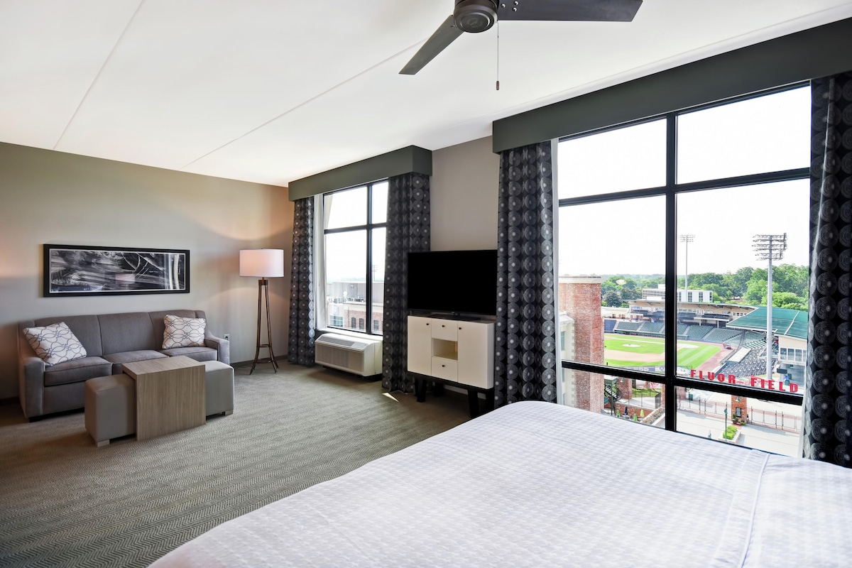 Een hotelkamer met een bed, een bank, een televisie en een groot raam met uitzicht op een honkbalveld.