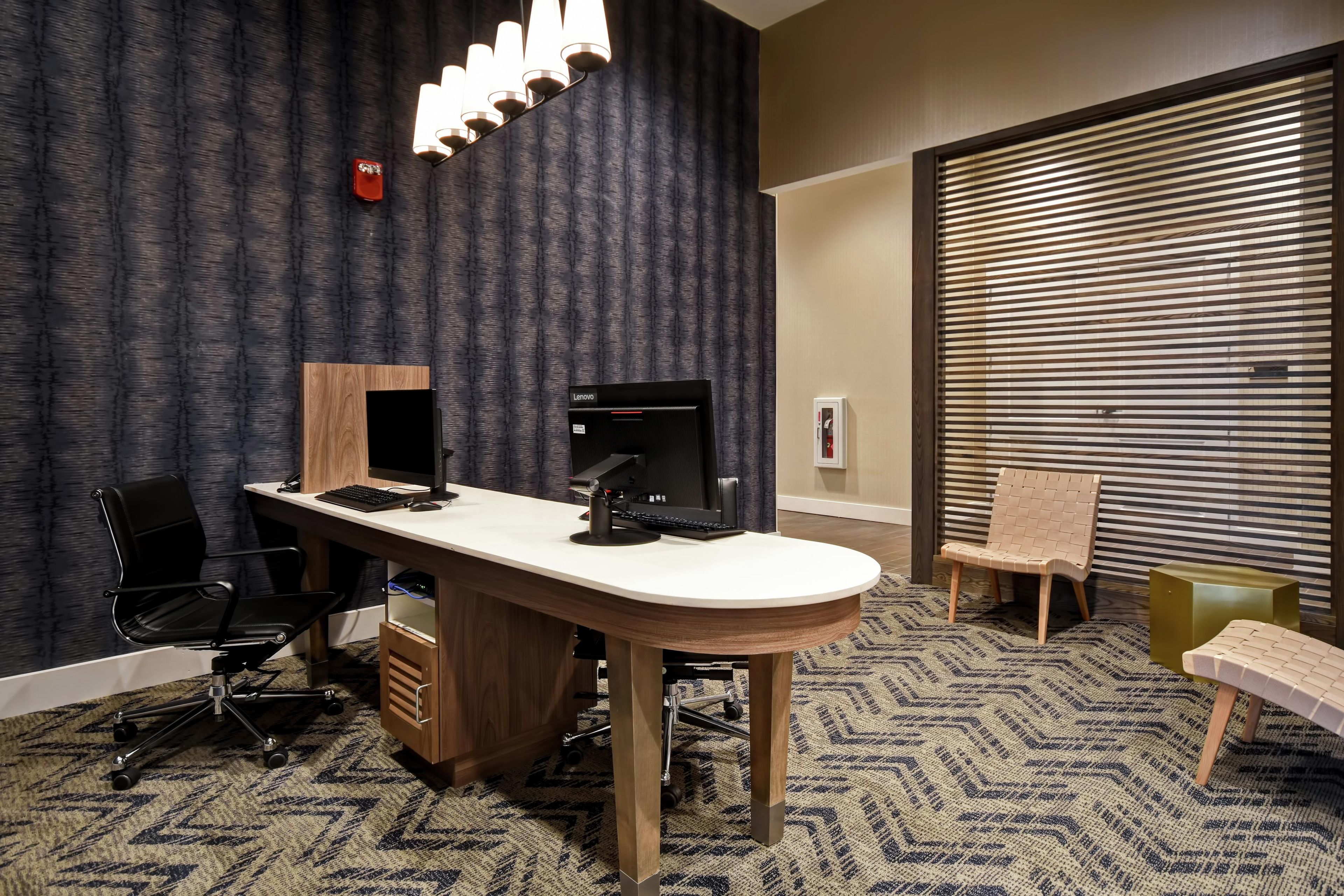 A hotel room with a desk, two chairs, a computer, and a large window with blinds.