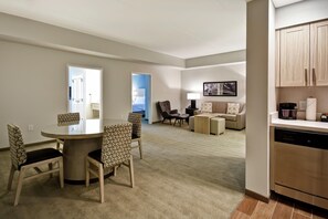 A modern living room with a dining area, a kitchen, and a bedroom visible through an open door. - Homewood Suites by Hilton Greenville Downtown (Greenville)
