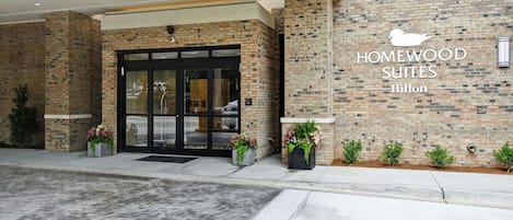 A hotel entrance with a brick facade, a glass door entrance, and the name "HOMEWOOD SUITES Hilton" displayed on the wall.