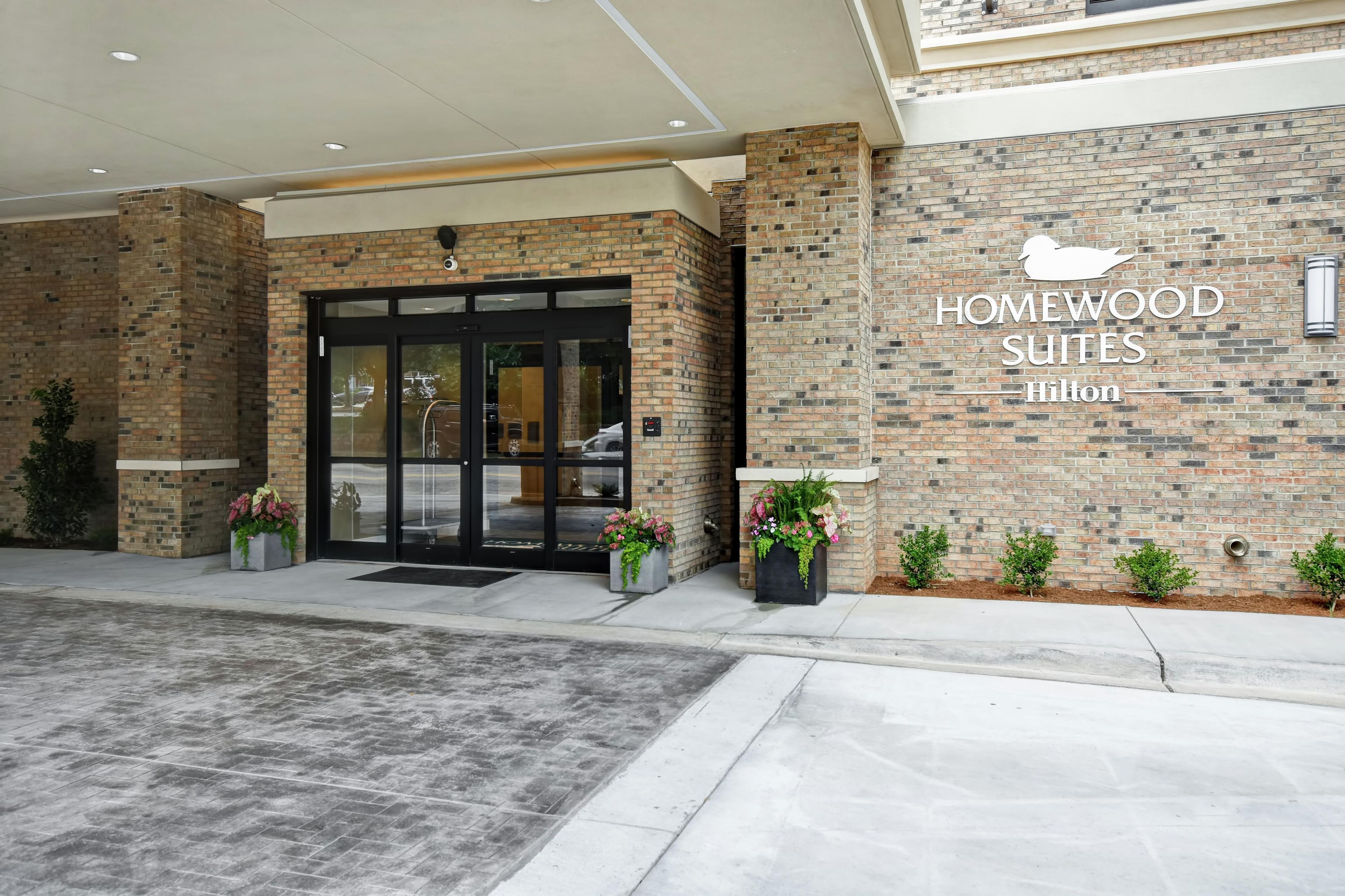 Entrada de un hotel con fachada de ladrillo, puerta de acceso de vidrio y un letrero que dice 'Homewood Suites Hilton'.