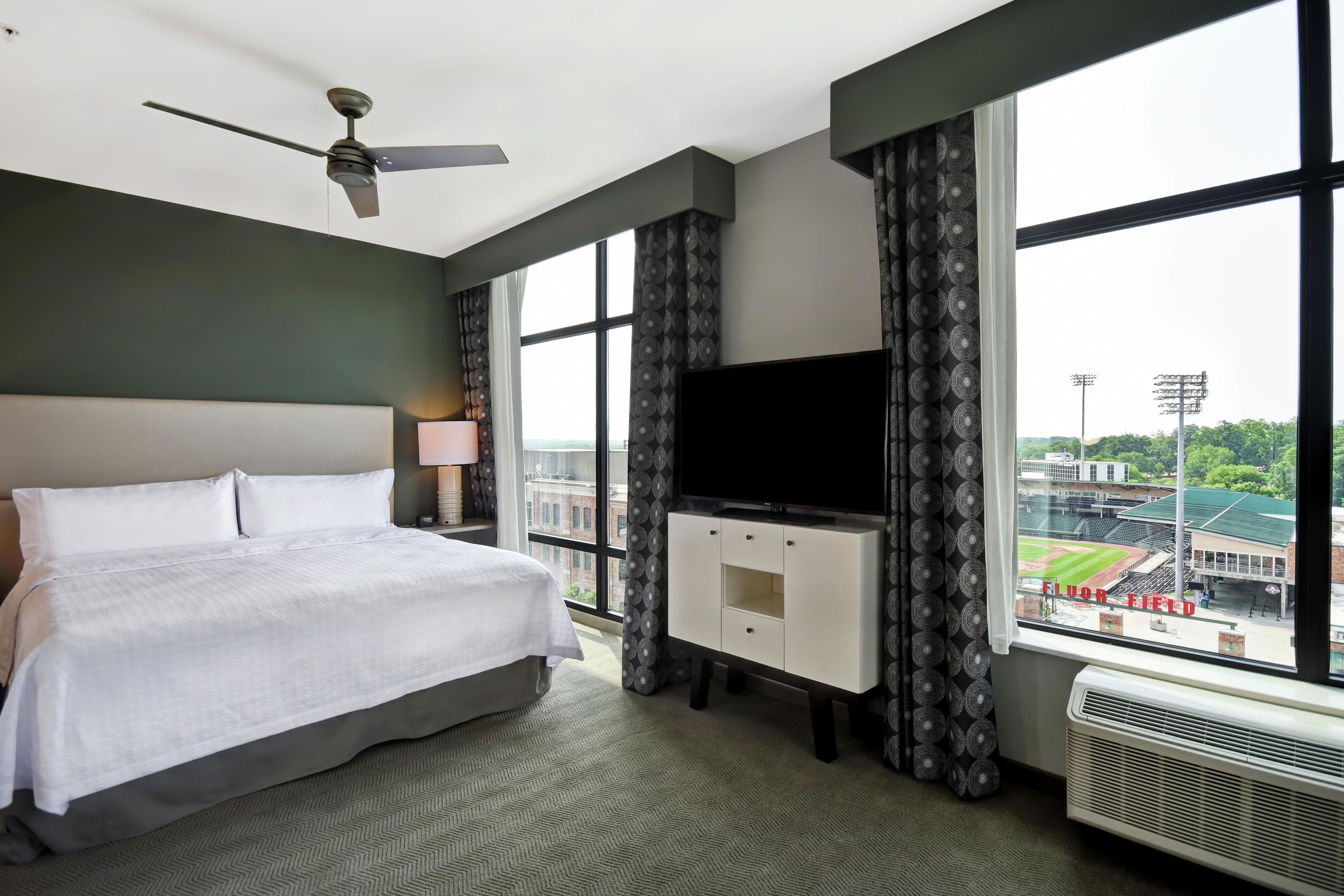 A hotel room with a bed, a flat-screen TV, a ceiling fan, and a large window offering a view of a stadium.