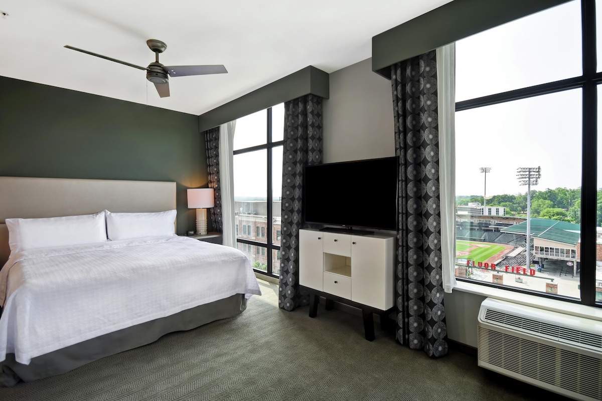 Een hotelkamer met een bed, een flatscreen televisie, een plafondventilator en een groot raam met uitzicht op een stadion.